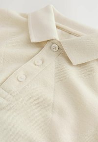 Beige poloshirt lavet af tekstureret stof, med klassisk krave, tre-knaps lukning og synlige strikkemønsterdetaljer.