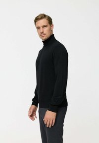 Fekete garbópulóver, puha anyagból készült, hosszú ujjaival, testhezálló fazonnal és bordázott mandzsettákkal. Szürke nadrággal viselve.