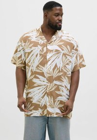 Chemise à manches courtes, boutonnée, affichant un fond marron avec des motifs de feuilles blanches. Le tissu semble lisse avec une coupe décontractée et un col.
