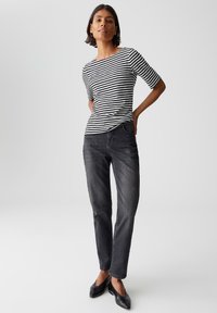 Femme se tenant avec les mains derrière le dos, portant une chemise à rayures noires et blanches, un jeans foncé et des ballerines noires pointues sur un fond neutre.