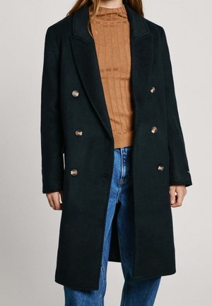 Femme portant un manteau noir croisé sur un pull côtelé marron et un jean bleu sur un fond uni.