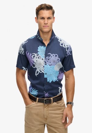 Camisa de manga corta azul marino con diseño floral en azul, púrpura y plateado. Cuenta con un cuello abotonado y una textura de tela suave.