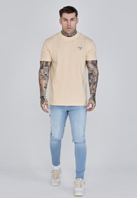 Világos bézs, rövid ujjú póló, kerek nyakkal és diszkrét logóval, világoskék skinny farmernadrággal és texturált fűzőjű fehér sportcipővel párosítva.