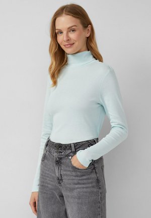 Kobieta z jasnobrązowymi włosami, ubrana w jasnoniebieski sweter z golfem i wysokim stanem, szare dżinsy, pozująca z jedną ręką w kieszeni na jednolitym tle.