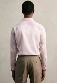 Chemise rose clair à manches longues avec un col classique, associée à un pantalon beige avec deux poches arrière et des accents discrets en métal.