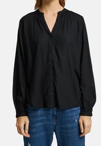 Sort langærmet bluse med V-hals, knaplukning foran og samlet detalj ved halsudskæringen. Fremstillet af glat stof, parret med blå jeans.