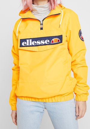Personne portant une veste à capuche ellesse jaune avec une poche zippée à l'avant et un jean bleu clair sur un fond uni.
