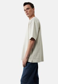 Jasno beżowy oversize'owy t-shirt z miękkiego materiału, z krótkimi rękawami, prostym dołem z bocznymi rozcięciami oraz luźnym krojem.