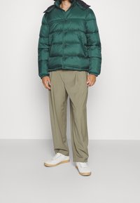 Vadderad grön jacka med kontrasterande svart krage och knappar, i kombination med beige vida byxor och vita sneakers på en enfärgad bakgrund.