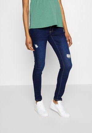 Kvinde iført mørkeblå, revnede skinny jeans, grøn top og hvide sneakers, står mod en simpel lys baggrund.