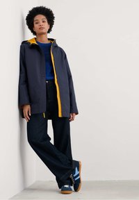 Jūrininkų lietpaltis su geltonu užtrauktuku ir gobtuvu, derinamas su mėlynu megztiniu ir tamsiai džinsinėmis plačiomis kelnėmis. Mėlyni ir rudi sportbačiai užbaigia įvaizdį.