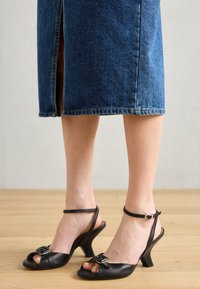 Black leather high-heeled sandals with open toes, adjustable ankle strap, and a decorative buckle. Curved heel and smooth texture.