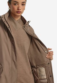 Brun vattentät jacka med dragkedja och tryckknappar. Inuti finns en mjuk beige foder och tvättrådsetikett. Texturerad tyg, avslappnad passform.