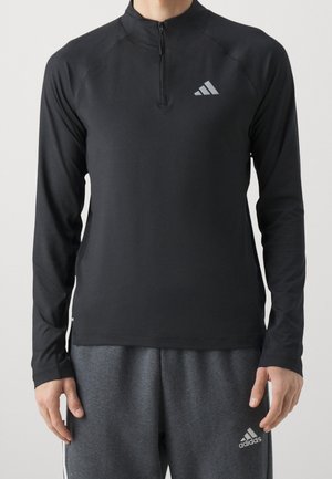 Homme portant un haut noir à manches longues avec fermeture éclair jusqu'au quart et un pantalon de survêtement gris Adidas avec logos sur la poitrine et la cuisse.