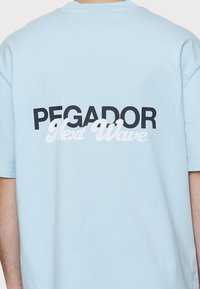 Světle modré bavlněné tričko s černým a bílým textem na zádech: "PEGADOR Next Wave." Krátké rukávy, kulatý výstřih, volný střih.