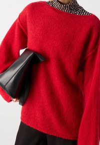 Persona che indossa un maglione lanoso rosso brillante sopra un dolcevita a righe zebrate, mentre tiene una borsa a soffietto in pelle nera piegata sotto un braccio.