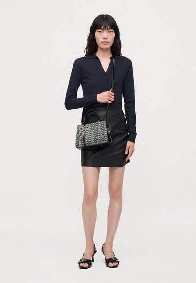 Une femme porte un haut bleu marine à manches longues, une minijupe en cuir noir, des sandales à talons et un sac à bandoulière DKNY à motifs, sur un fond uni.
