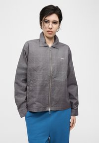 Άτομο με κοντά σκούρα μαλλιά και γυαλιά, που φοράει γκρι καρό ζακέτα με φερμουάρ και μπλε παντελόνι, στέκεται μπροστά σε λευκό φόντο.