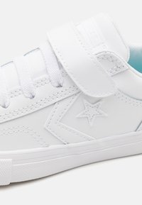 Sneaker blanche en matériau synthétique, dotée d'une sangle velcro et de lacets, avec des détails cousus et un emblème en forme d'étoile sur le côté.