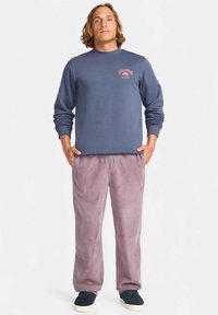 Sudadera azul marino con puños acanalados, que presenta un pequeño logo rosa. Pantalones de pana color lila claro con bolsillos laterales y cintura elástica. Zapatillas negras.