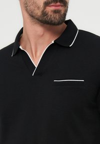 Κοντινό πλάνο ενός άνδρα που φοράει μαύρο πόλο πουκάμισο με λευκή περιμετρική ραφή στον γιακά και στην άκρη της τσέπης στο στήθος, εστιάζοντας στη γραμμή του λαιμού και στα γένια.