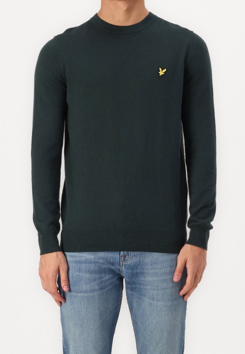 Jersey de punto verde oscuro con mangas largas, cuello redondo y puños de canalé. Presenta un pequeño logo amarillo en el pecho. Combinado con unos vaqueros azul claro.