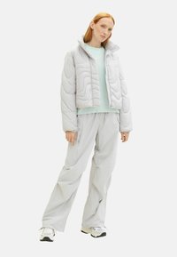 Jachetă puffer matlasată de culoare gri deschis, cu un guler înalt, asociată cu un pulover verde mentă și pantaloni gri largi. Prezintă o textură netedă.