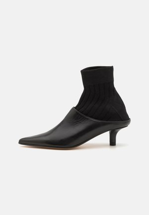 Schwarzer Lederschuh mit spitzem Zeh und geripptem schwarzem Strickansatz am Knöchel. Verfügt über einen niedrigen, zylindrischen Absatz und eine glatte Oberfläche.