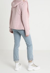 Giacca in pile rosa chiaro con cappuccio, abbinata a jeans azzurri e stivaletti alla caviglia con suola spessa bianchi. Tessuto strutturato e design casual.