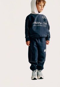 Mørkeblå sweatshirt med hvidt logo tekst og snøre i hætten, kombineret med matchende joggingbukser med kontrasterende syningsdetaljer og lommer.