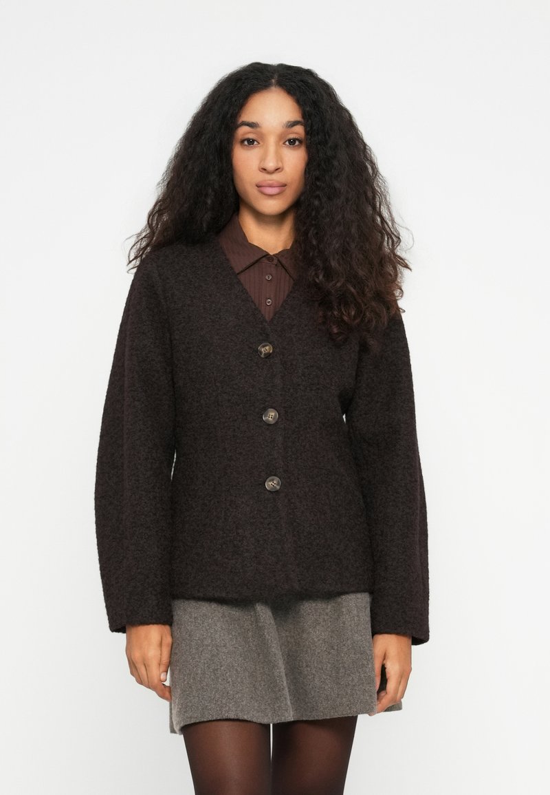 Vrouw met krullend haar, gekleed in een donkerbruine gebuttonde cardigan over een bruine blouse met kraag en een grijze rok met zwarte panty, tegen een witte achtergrond.