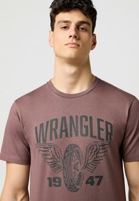 Brun bomull T-shirt med en svart grafik av vingar och ett däck med texten 'WRANGLER 1947' i fet stil, korta ärmar och rund halsringning.