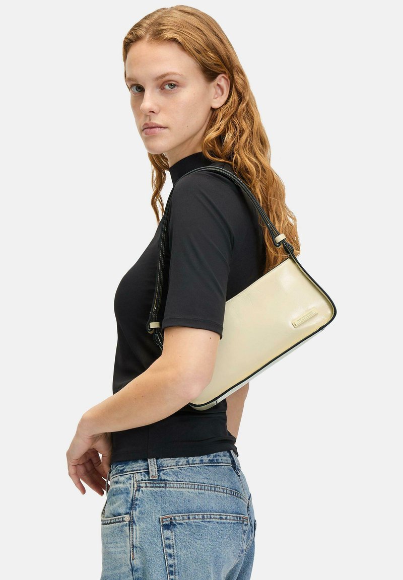 Borsa a tracolla crema con rifiniture nere, forma rettangolare, materiale texturizzato e tracolla regolabile, tenuta contro una maglietta nera e jeans blu.