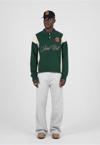 Polo verde e crema a maniche lunghe con logo sul petto, abbinato a pantaloni grigi e pantofole. Cappellino nero con logo ricamato.