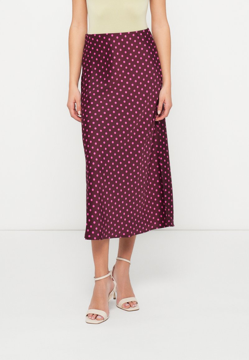 Uma saia midi cor borgonha com uma textura brilhante, apresentando bolinhas cor-de-rosa por toda a parte. Usada com sapatos de salto bejes com tiras.