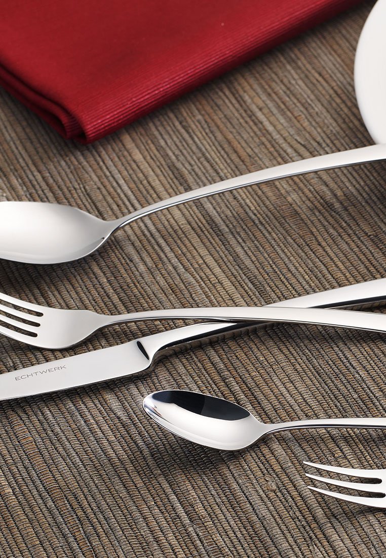 Juego de cubiertos de acero inoxidable que incluye un cuchillo, un tenedor y una cuchara con un acabado pulido y elegante sobre un mantel texturizado, junto a una servilleta de tela roja.