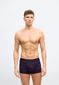 Άνδρας χωρίς πουκάμισο με μπλε σκούρα ανδρικά εσώρουχα boxer με κόκκινη καρδιά και σχέδιο κροκοδείλου, στέκεται μπροστά σε απλό λευκό φόντο.