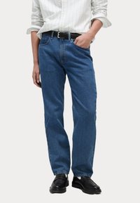 Vaqueros de mezclilla azul con corte recto, con bolsillos laterales y un cinturón. Llevados con una camisa a rayas y zapatos negros.