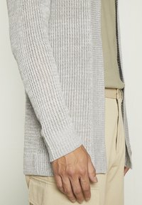 Helles grauer Strickcardigan mit geripptem Muster, offenem Frontbereich und langen Ärmeln, kombiniert mit einem beigen Oberteil und cremefarbenen Hosen.