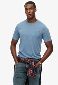 Camiseta de algodón azul con cuello redondo y logotipo en relieve. Llevada con jeans oscuros y una camisa de cuadros atada a la cintura. Cinturón y reloj marrones.