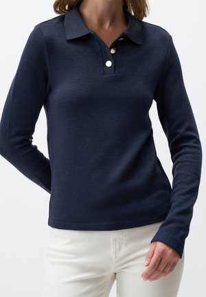 Mujer con camisa polo de punto azul marino de manga larga con dos botones plateados, combinada con pantalones blancos, posando frente a un fondo liso.