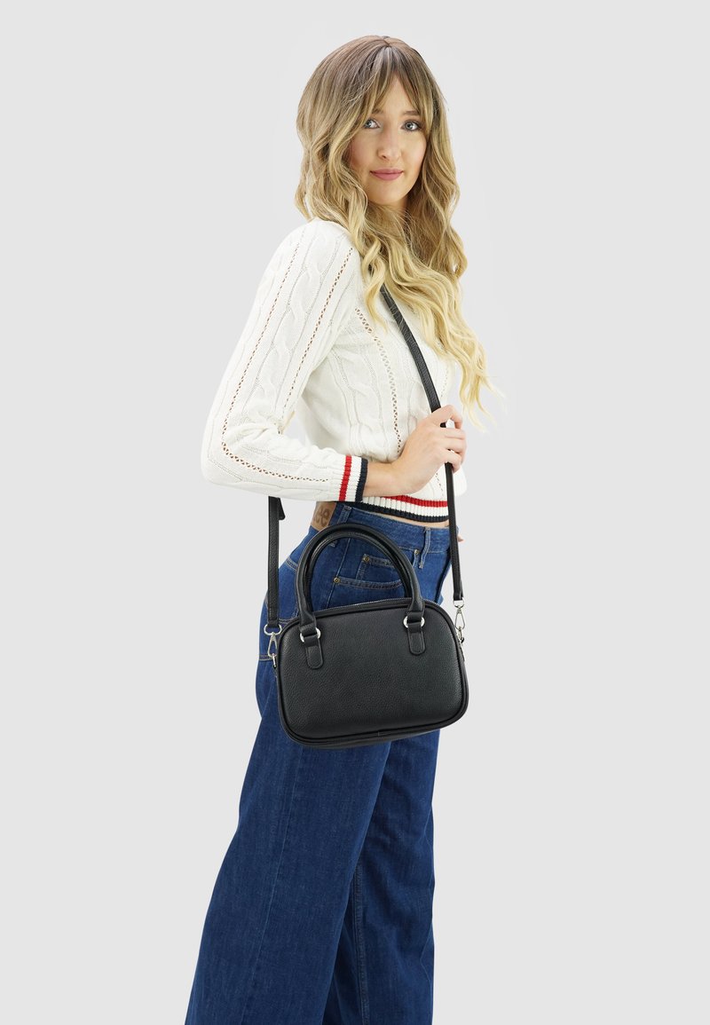 Vrouw met lang blond haar, gekleed in een witte gebreide trui en blauwe jeans, houdt een zwarte leren schoudertas vast tegen een grijze achtergrond.