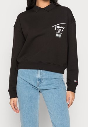 Czarna bluza polo z długim rękawem z białym logo Tommy i nazwami miast na piersi, w połączeniu z jasnoniebieskimi dżinsami na stojącej osobie.