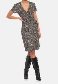 Žena ve černých šatech s krátkým rukávem a bílým květinovým vzorem, s černými koženými botami po kolena, stojící s rukou na bok.