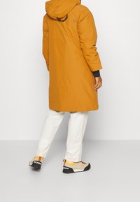 Vandafvisende, orange parka med hætte, der har sorte detaljer og et logo. Kombineret med hvide bukser og multikolorede outdoor sko.
