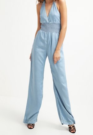 Jasnoniebieski dżinsowy kombinezon z dekoltem na halter, marszczonym stanem, szerokimi nogawkami i subtelnym połyskiem. Noszony z czarnymi sandałami na paski.