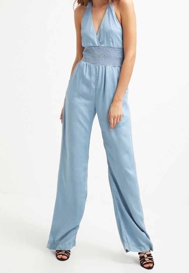 Jumpsuit en denim bleu clair avec un col halter, une taille smockée, un design à jambes larges et un léger éclat. Porté avec des sandales noires à lanières.