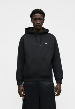 Czarna bluza z kapturem z logo Adidas na piersi, noszona przez stojącą osobę na jednolitym, jasnym tle.