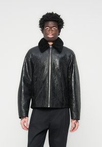 Veste en faux cuir noir avec une finition texturée, dotée d'un grand col en fourrure noire, d'une fermeture zippée et de deux poches latérales.