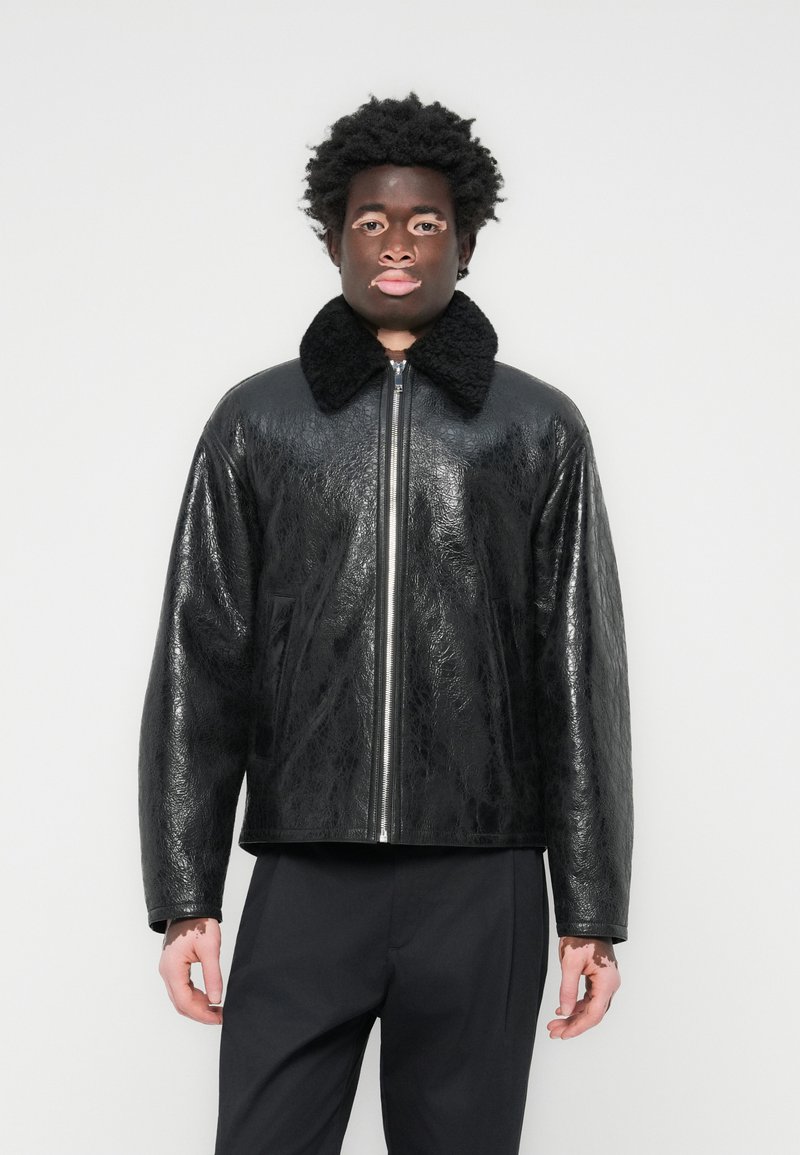 Veste en faux cuir noir avec une finition texturée, dotée d'un grand col en fourrure noire, d'une fermeture zippée et de deux poches latérales.